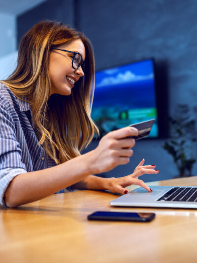 Side view of young charming caucasian woman with eyeglasses holding credit card and using laptop for online shopping while sitting at dining table at home.