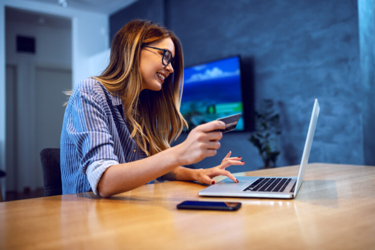 Side view of young charming caucasian woman with eyeglasses holding credit card and using laptop for online shopping while sitting at dining table at home.